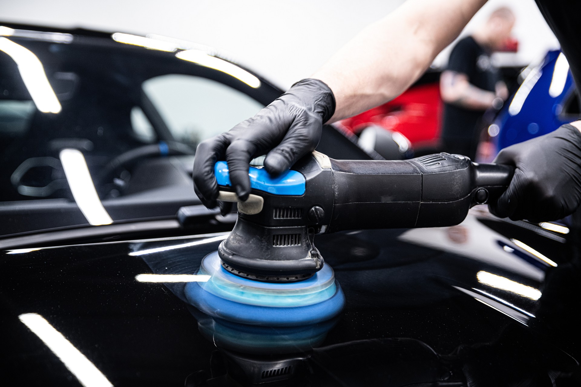 Mechanic wearing black gloves polishing car paint with a machine in a detailing studio.